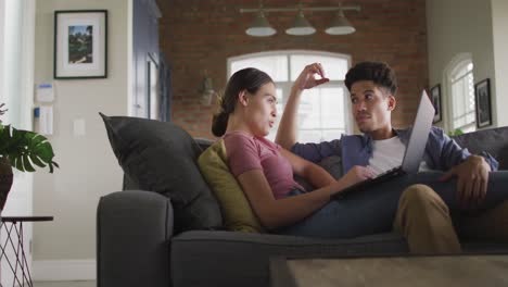 Happy-biracial-couple-sitting-on-sofa-in-living-room,-using-laptop