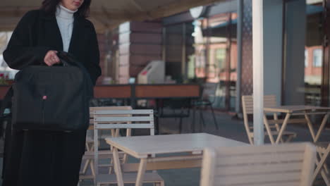 young investor in black coat relaxes at a cafeteria, setting her bag on the table against a blurred background with urban elements like poles, flowers, and buildings