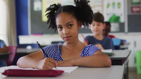 Portrait-of-happy-mixed-race-schoolgirl-sitting-at-classroom,-making-notes,-looking-at-camera