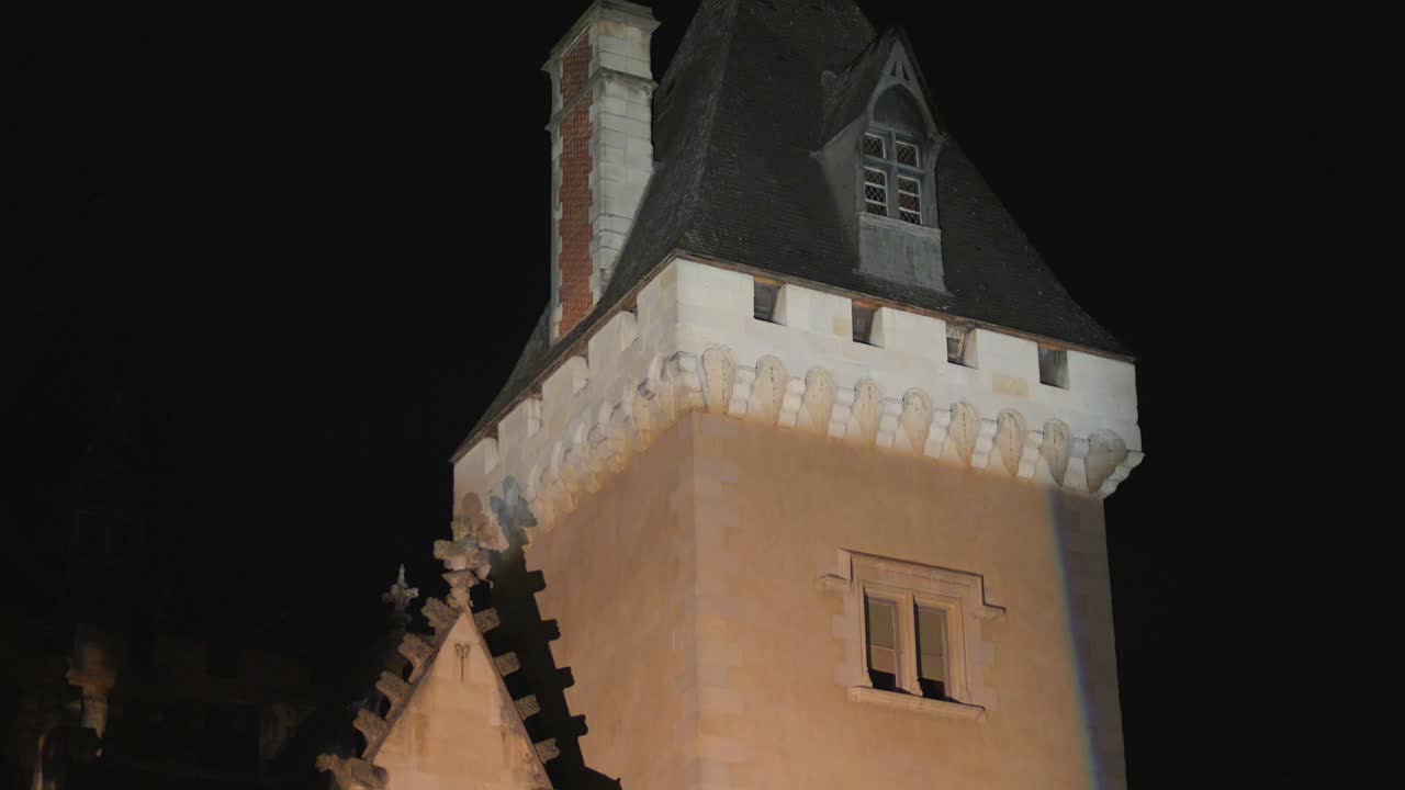 A dramatic close-up of a medieval stone tower at the Château de Pau in France, illuminated against the dark night sky. The shot captures the historic architecture and atmosphere
