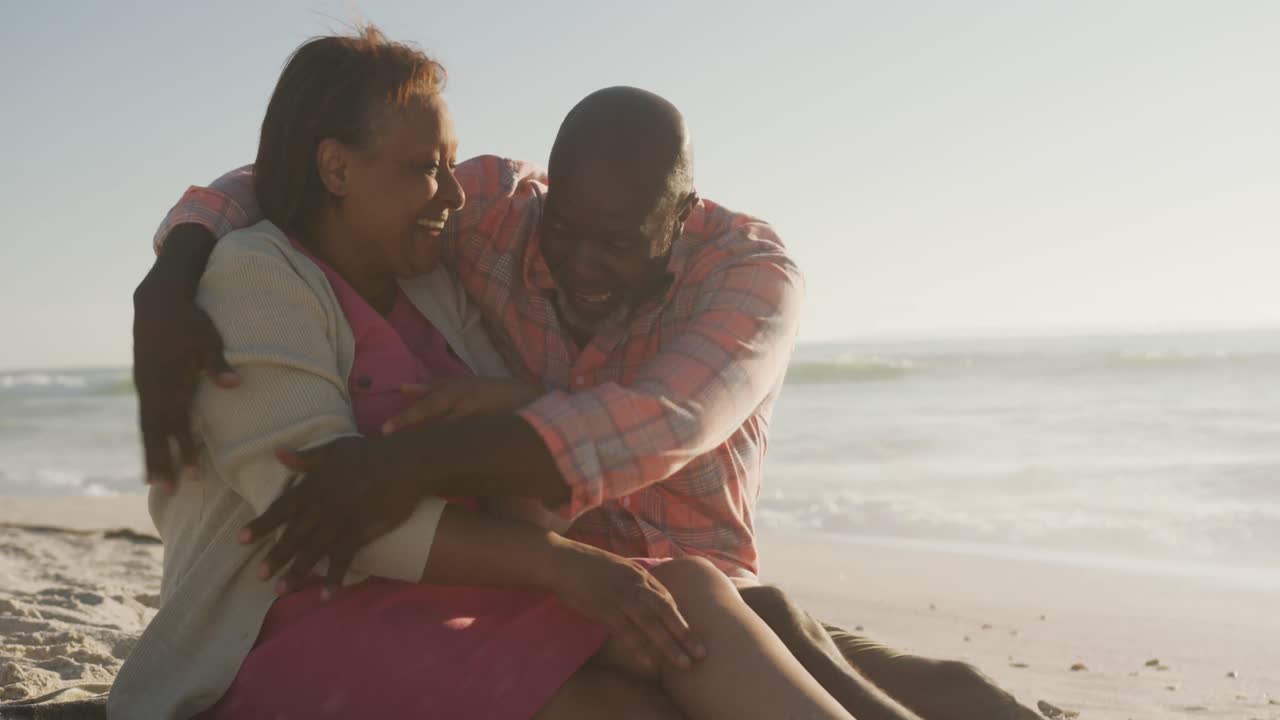 una pareja afroamericana de alto nivel sonriente abrazándose y sentada en una playa soleada