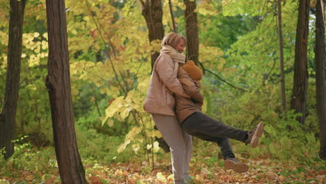 joyful mother spinning child in golden autumn forest, both dressed warmly in jackets and beanies, surrounded by sunlit yellow leaves and vibrant trees, sharing playful moment