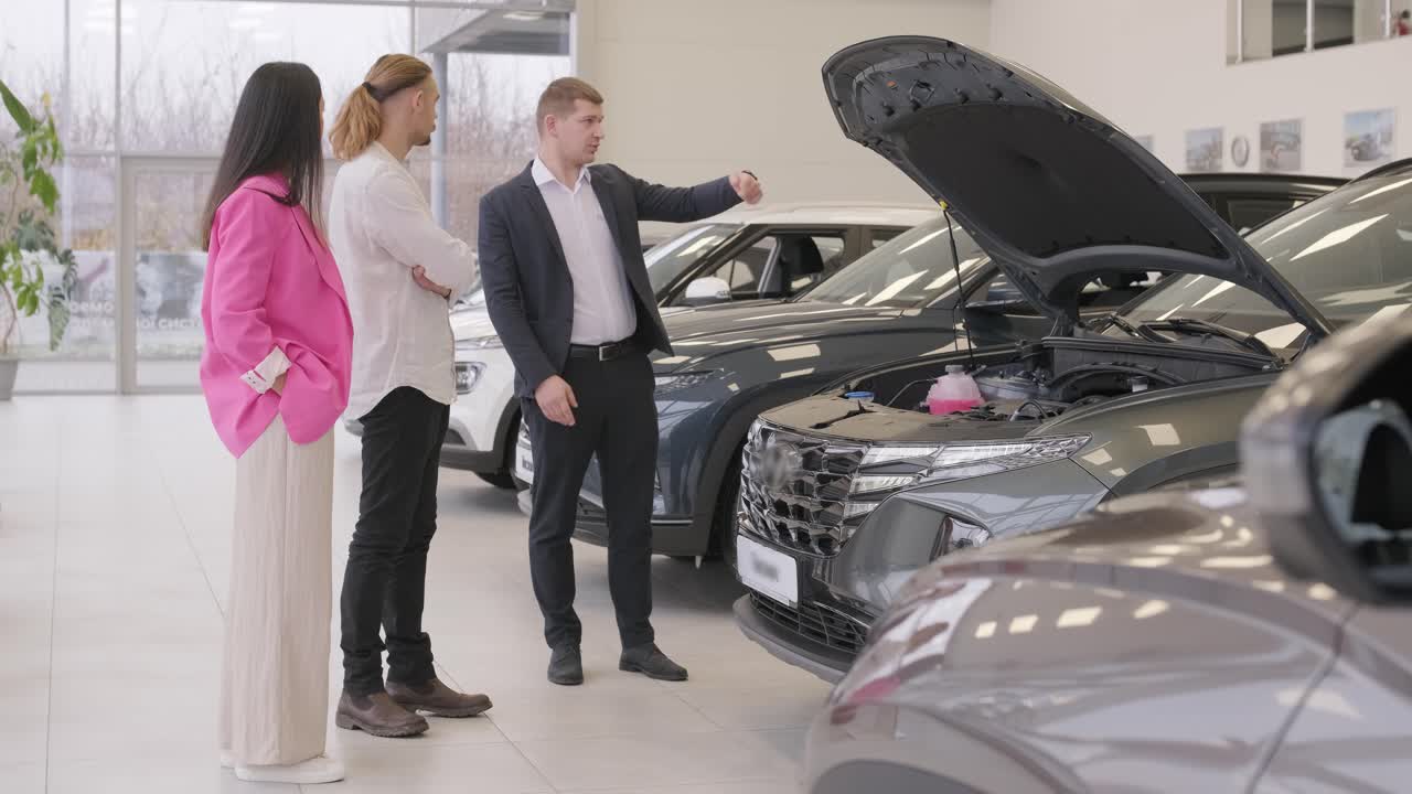 젊은 자동차 판매자가 대리점 살롱에서 젊은 부부에게 새로운 자동차를 보여주고 있습니다.