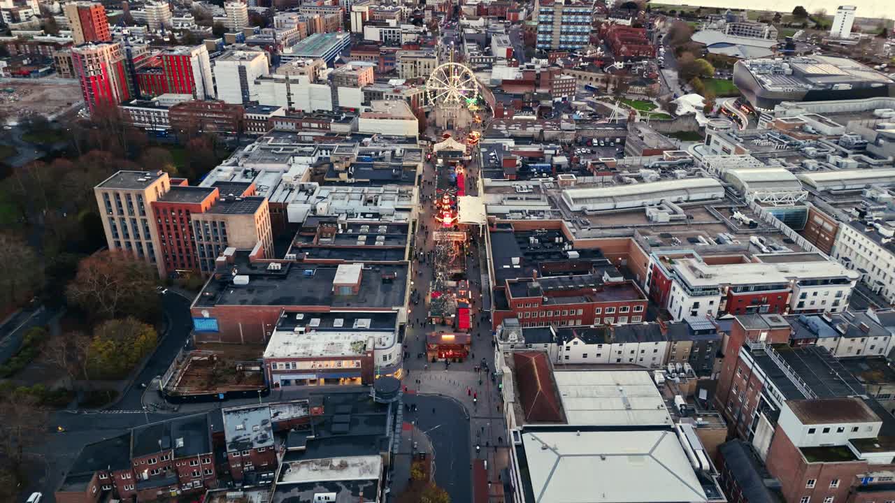 High-angle aerial drone shot over Southampton’s Christmas Market at sunset, capturing glowing festive stalls, warm lights, and steady crowds weaving through the square as the city softens at winter