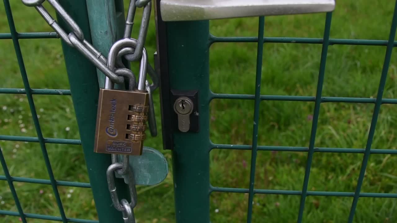 Close up panning up view of brown rectangular lock on silver chain on green metal gate.
