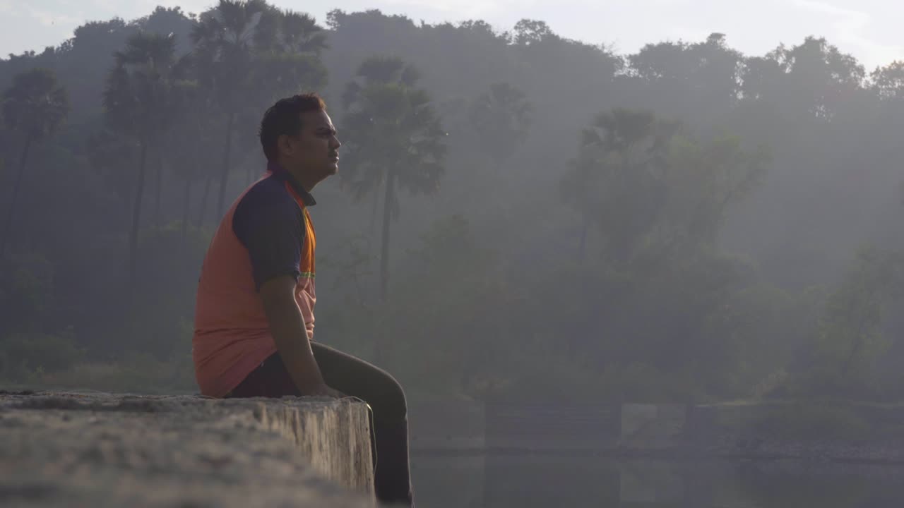 호수에 선착장에 앉아 있는 소년은 일출 아침에 경치를 편안하게 즐기고 있습니다.