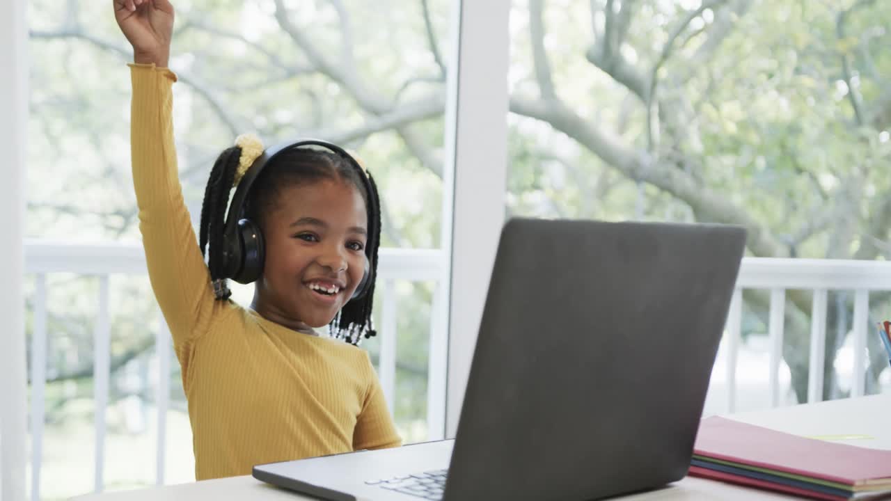 Happy african american girl using laptop and headphones raising hand in online class, slow motion