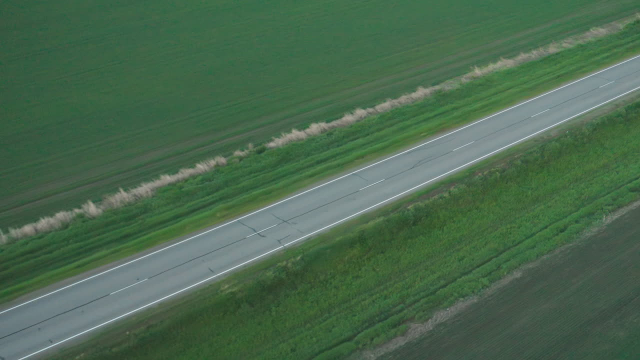 overhead drone view of straight two-lane road with crisp white markings cutting through lush green farmland under soft dusky light, highlighting contrast between asphalt ribbon