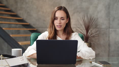 Joyful-business-woman-making-web-call-on-laptop-camera-in-modern-office.