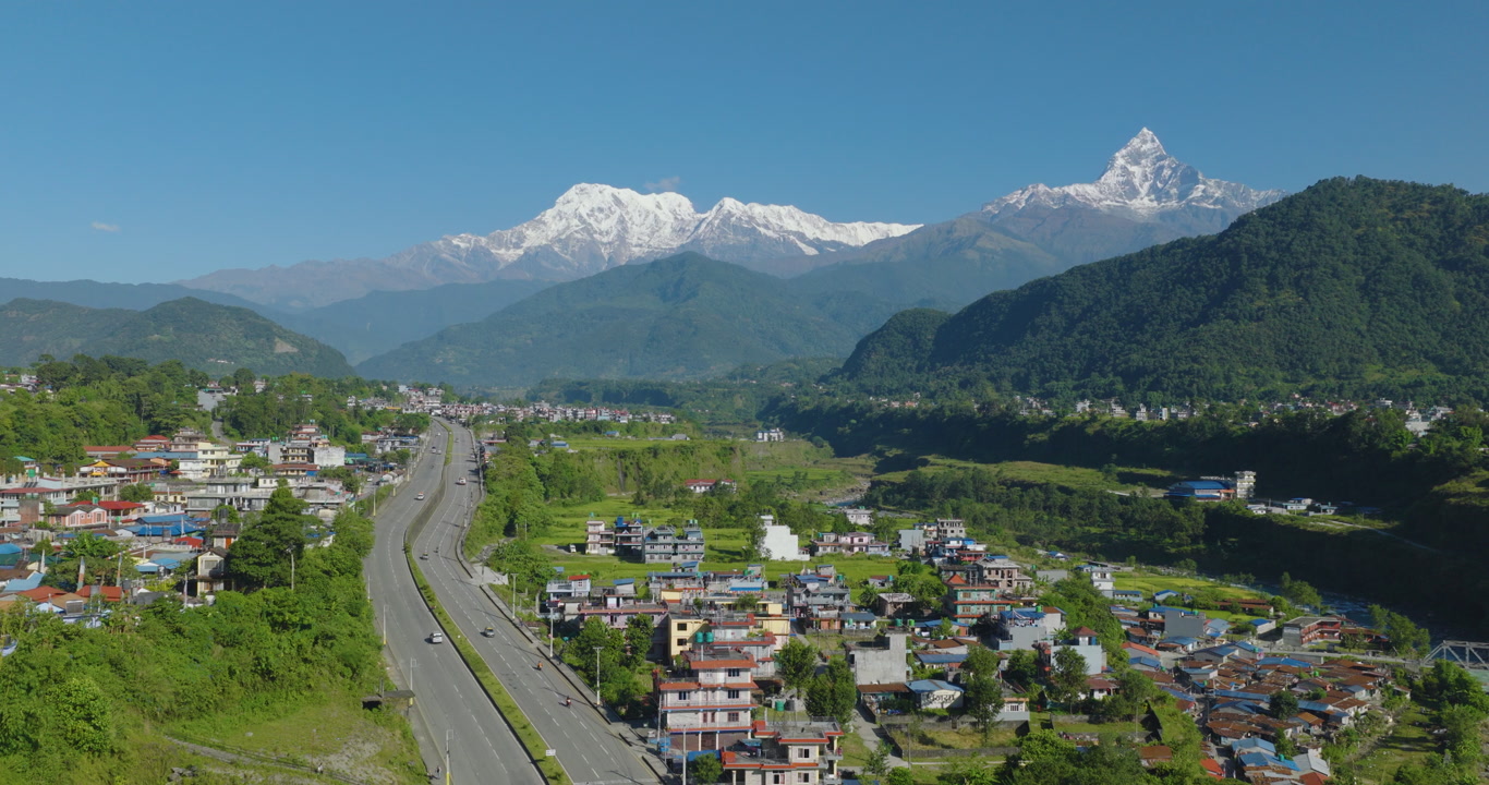 Aerial reveal shot of Prithvi Highway passing a village and green forest beside the Gandaki River with Annapurna and Machhapuchhre scenic mountain range rising in Pokhara Nepal.