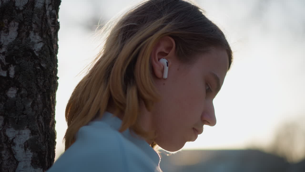 Un*adolescente bianco appoggiato a un albero che ascolta la musica, uno studente che si prende una pausa di riflessione durante un tramonto dorato, auricolari nelle orecchie, profilo retroilluminato delicato, stato d'animo calmo, sfondo naturale, zaino ai piedi