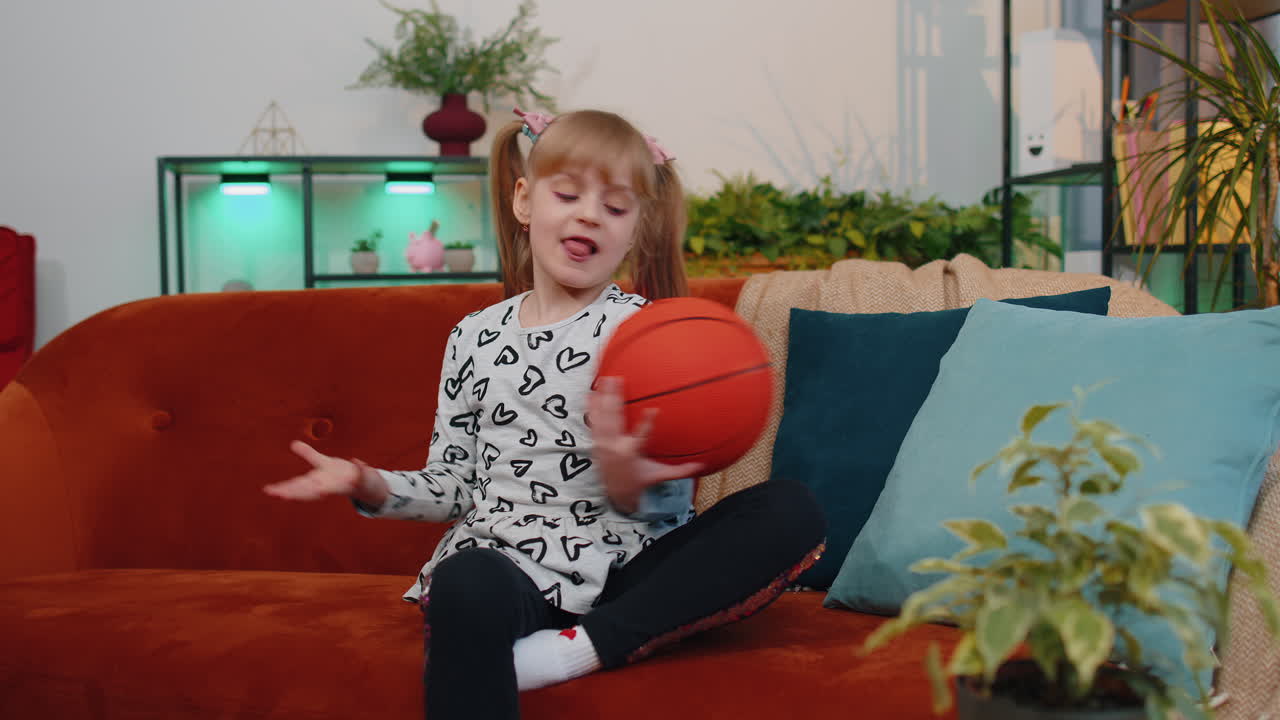 Young little children girl basketball fan holding ball looking at camera, training dribbling at home