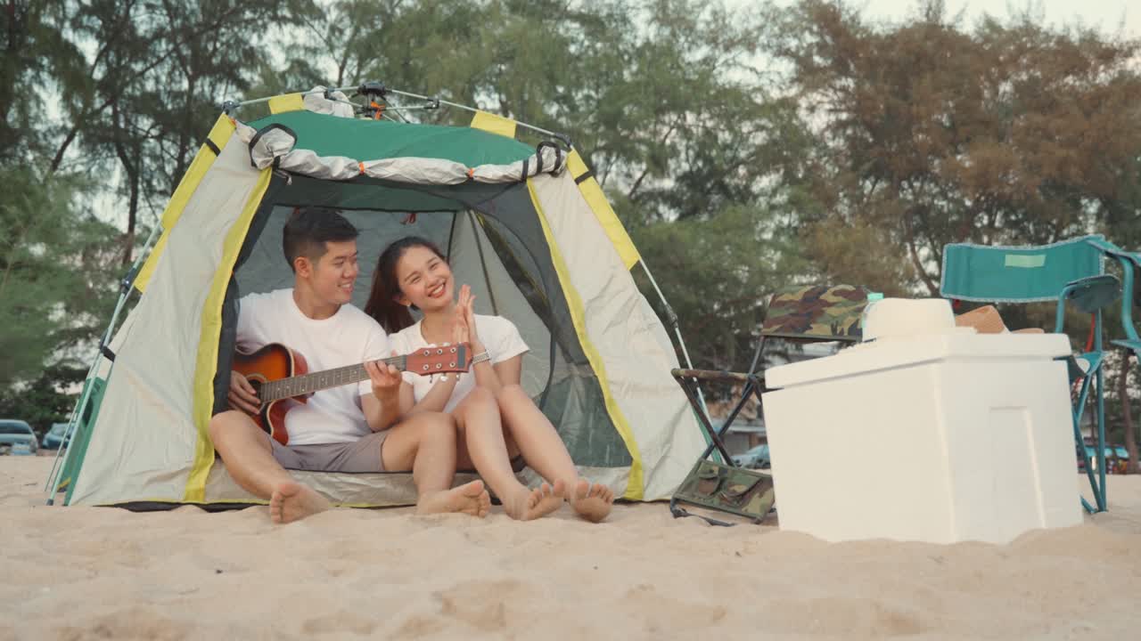 젊은 사랑하는 커플은 캠핑하는 동안 아름답게 기타를 연주하는 동안 해변에 캠핑 텐트에 앉아
