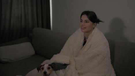 adult woman covered with blanket watching movie at home with dog pet jack russell terrier