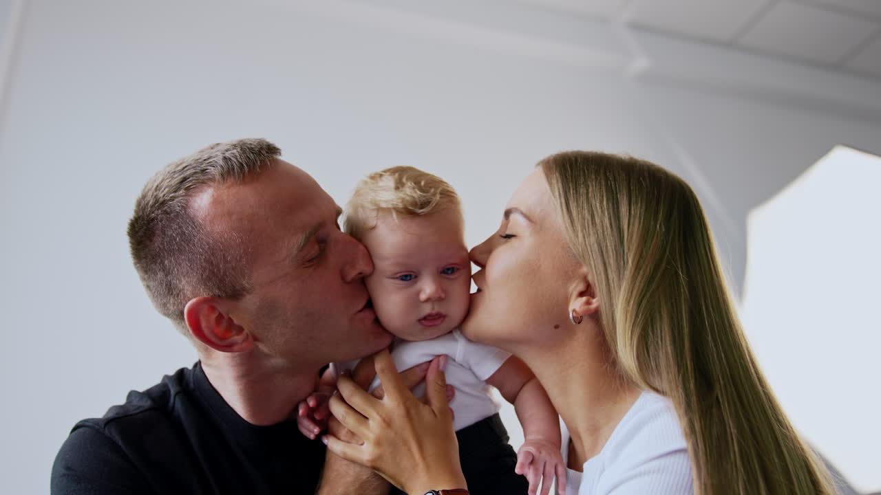 Adorable blond blue-eyed baby boy in hands of his parents. Loving parents kiss their beloved infant on the cheeks.