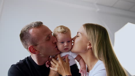 adorable blond blue-eyed baby boy in hands of his parents. loving parents kiss their beloved infant on the cheeks.