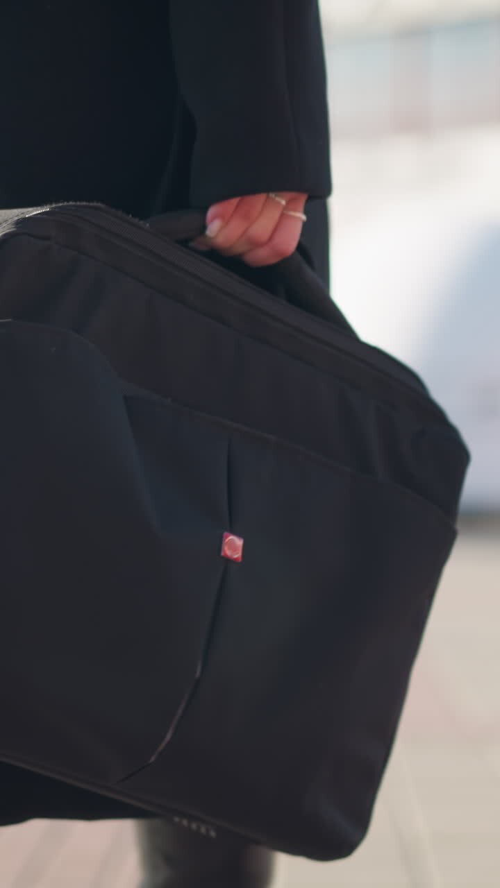 Close-up of black bag carried by woman in stylish black coat and leather boots walking confidently on sunny urban street, with blurred modern architecture and green bikes in the background