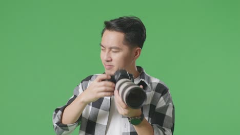 close up of asian photographer using a camera taking pictures and flexing his bicep while standing on green screen background in the studio