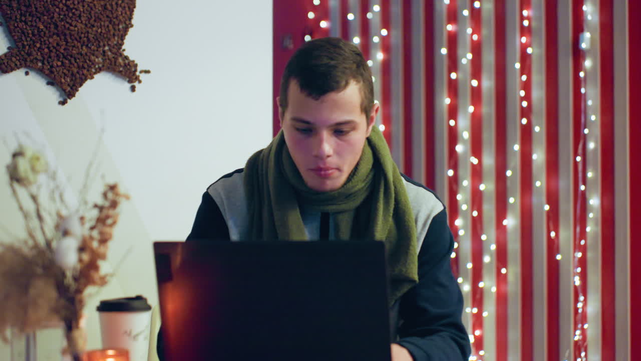 Caucasian man at laptop in cafe, latenight freelancer typing and researching, warm candle glow, dried flowers and string lights, wearing green scarf and jacket, concentrated expression, cozy ambient