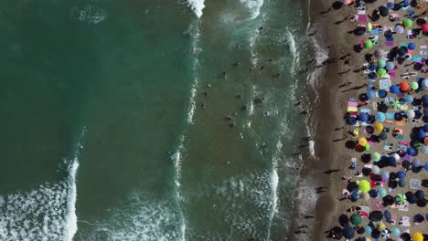 Vista-De-Pájaro-De-Personas-Nadando-En-Olas-En-Una-Playa-Concurrida-Con-Sombrillas