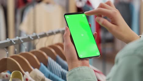 clothing store: female using smartphone with chroma key green screen display. clothes hanger with stylish branded items for retail sale in the background. close up footage of mobile device.