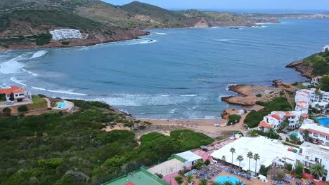 Wide-angle-aerial-view-of-people-on-vacation,-playing-tennis,-in-the-swimming-pool-and-relaxing-by-the-beach-in-Minorca,-Spain