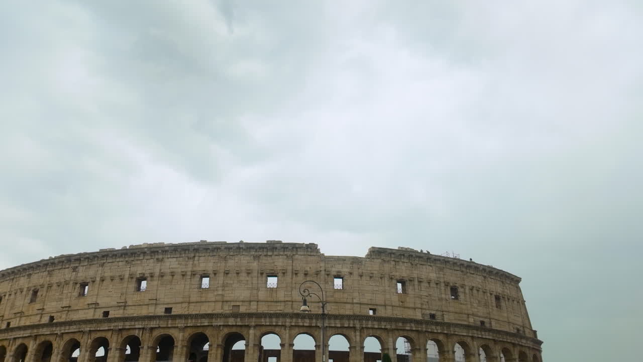 Upper section of the ancient Colosseum, with its iconic arches and a traditional street lamp, set against a cloudy sky