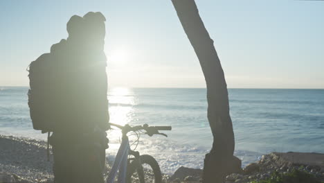 Der-Radfahrer-Kommt-Am-Kiesstrand-An,-Um-Einen-Spektakulären-Sonnenaufgang-Zu-Erleben,-Nimmt-Seinen-Rucksack-Ab-Und-Betrachtet-Die-Landschaft