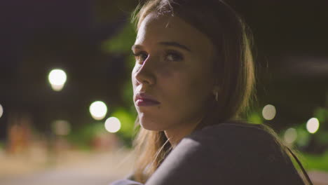 close-up of thoughtful young woman at night sitting outdoors, illuminated by soft park lighting with blurred background of glowing streetlights and greenery, expressing calm reflection