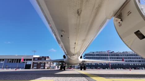 external view of the lower part of the fuselage of a dirty white color fuselage jet from the nose to thre rear part, tilting down, with some buildings at the back. 4k