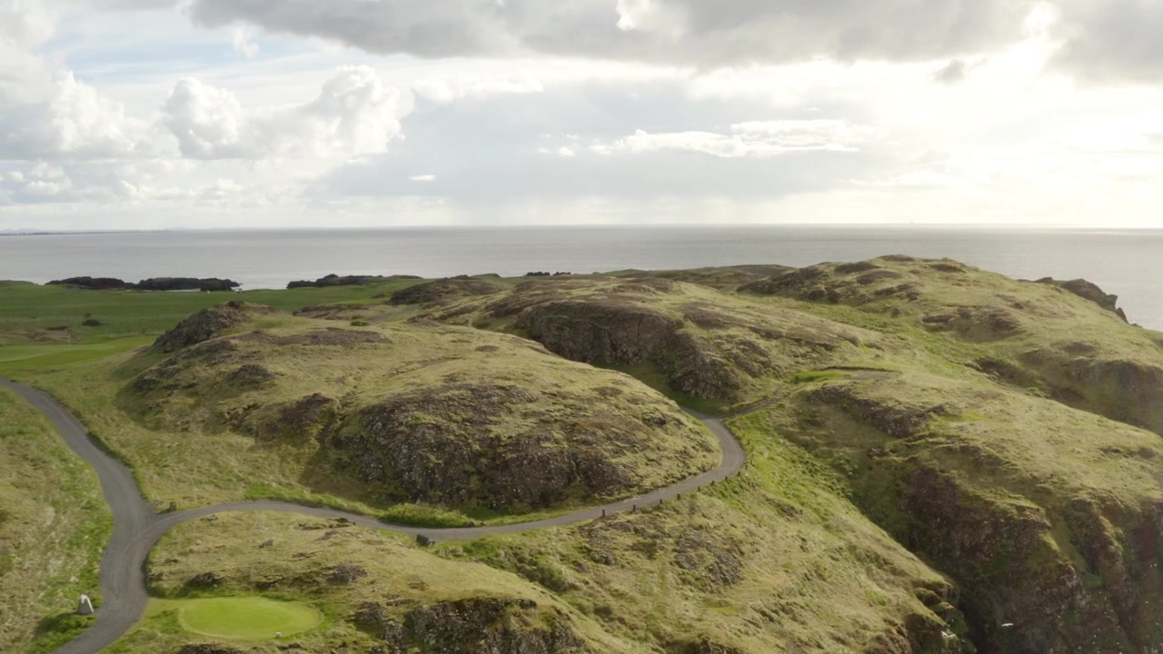 Luftfoto af en naturskøn kystgolfbane med grønne bakker og en snoet sti. En strandskade flyver forbi, mens to personer står ved en golfvogn.