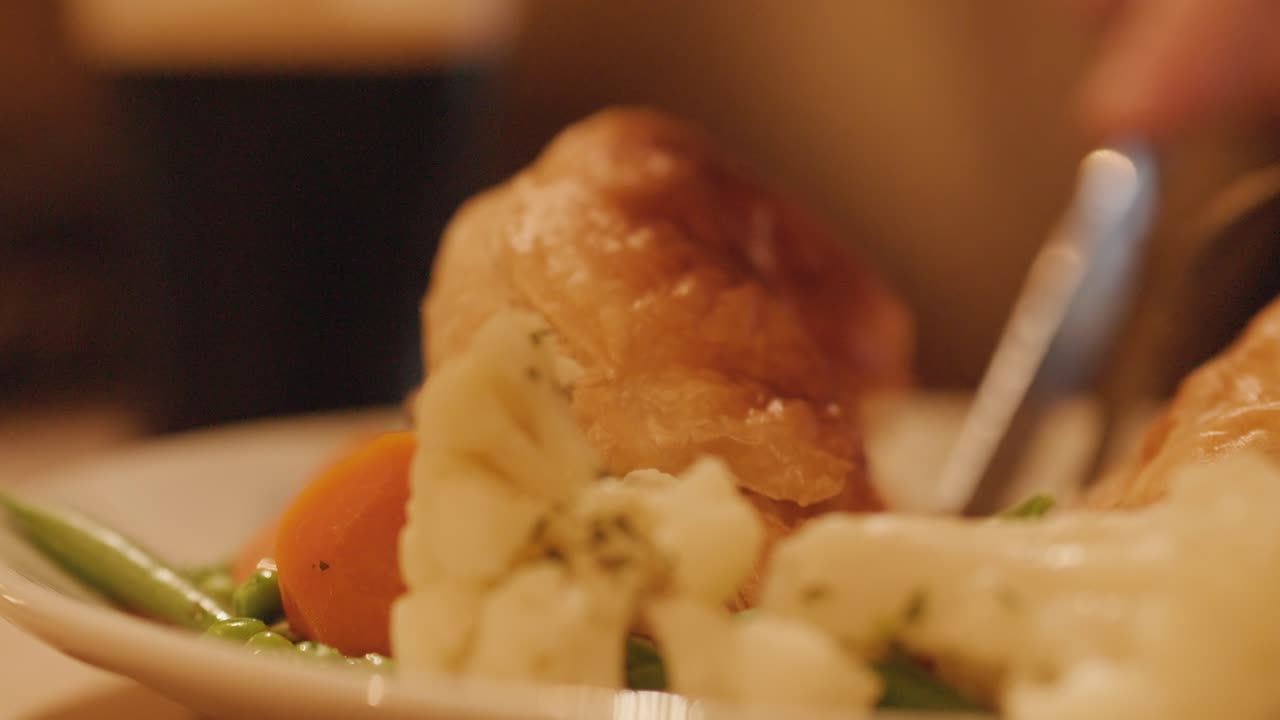 A hand slices into a golden steak pie served with vegetables and beer in a warmly lit pub, captured in close-up with shallow focus
