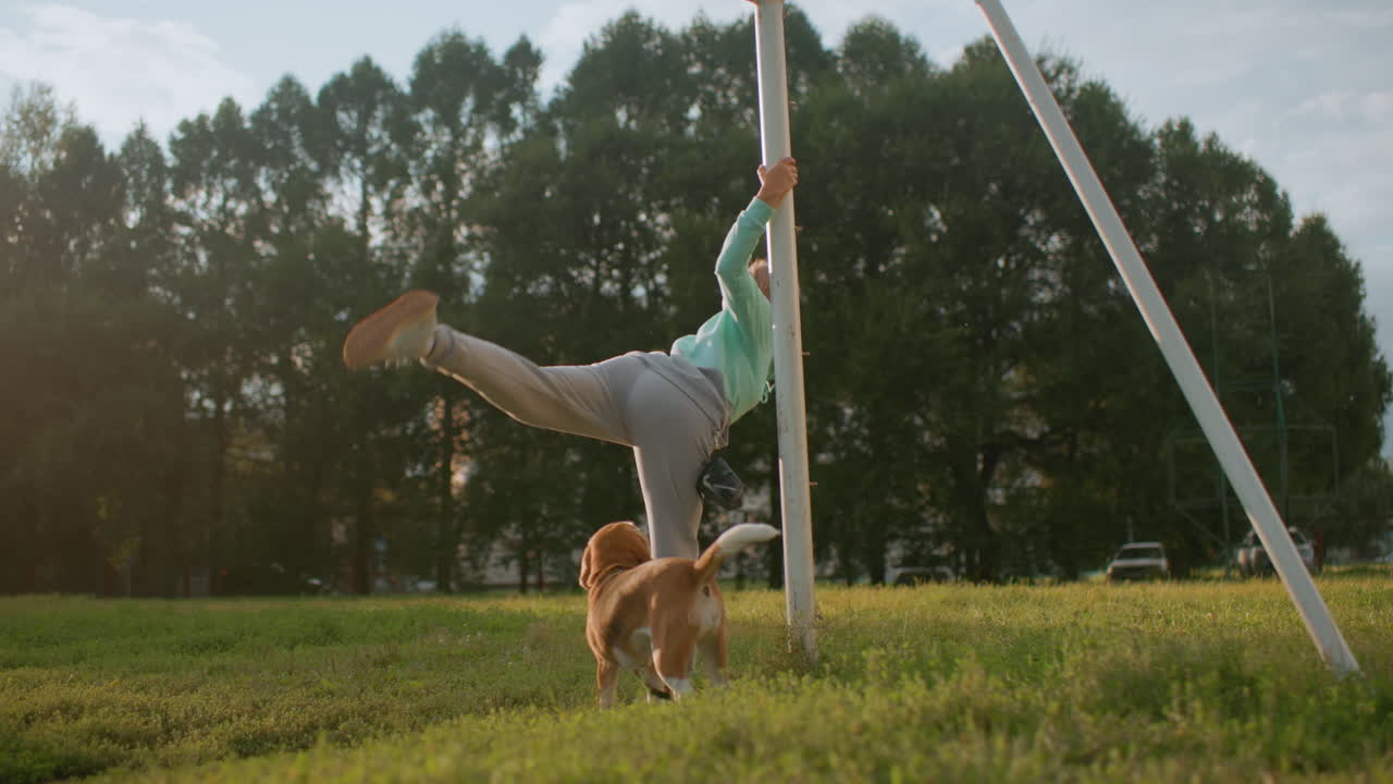 Coach holding stand performing body stretch workout outdoors on grassy field under sunny sky while dog roams nearby in park surrounded by trees capturing moment of fitness, balance, strength