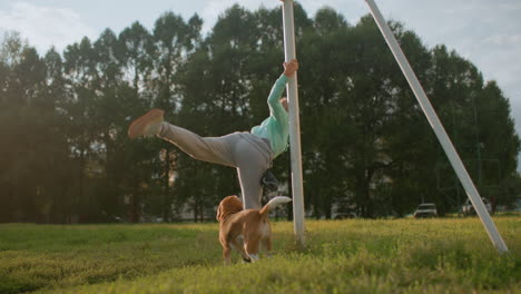 coach holding stand performing body stretch workout outdoors on grassy field under sunny sky while dog roams nearby in park surrounded by trees capturing moment of fitness, balance, strength