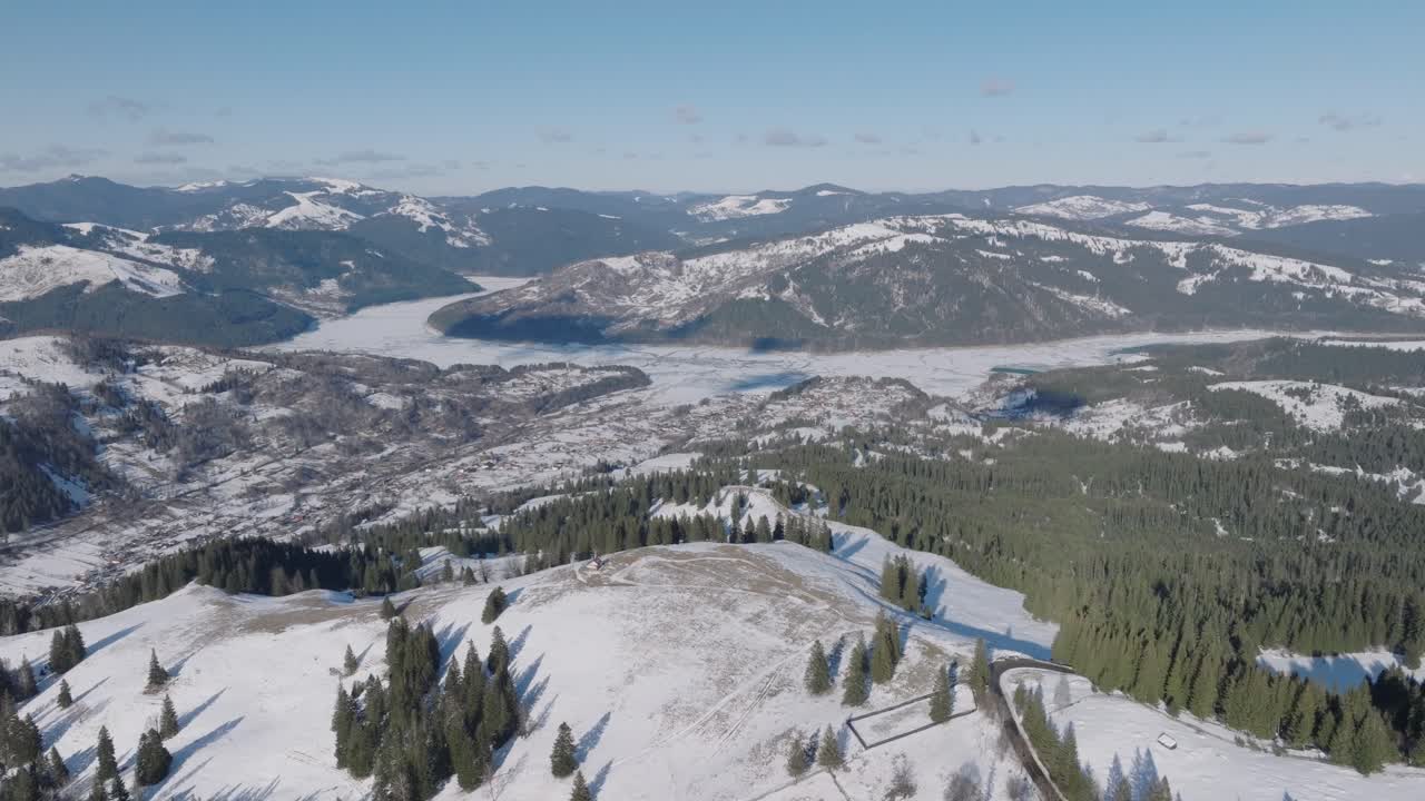 Snow-covered mountains and forest under bright blue sky with distant lake, aerial view