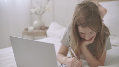 school girl is lying on bed with laptop and copybook writing homework and looking on display of laptop