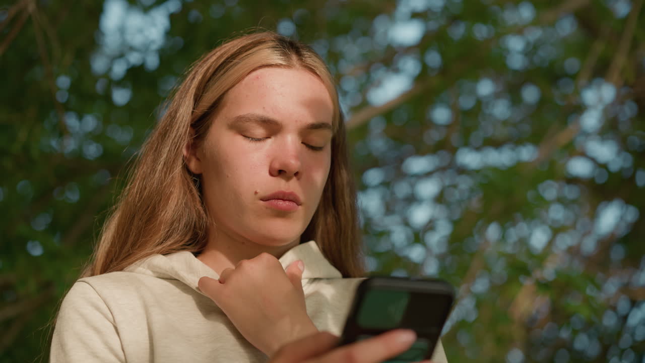 primer plano de una joven con una pequeña mancha facial usando el teléfono atentamente mientras sostiene su collar de capucha, la luz del sol iluminando suavemente su cara a través de las hojas de los árboles, con un fondo verde borroso