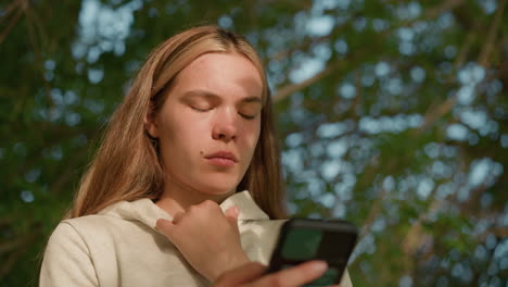close-up of young girl with small facial spot intently using phone while holding her hood collar, sunlight softly illuminating her face through tree leaves, with blurred green background