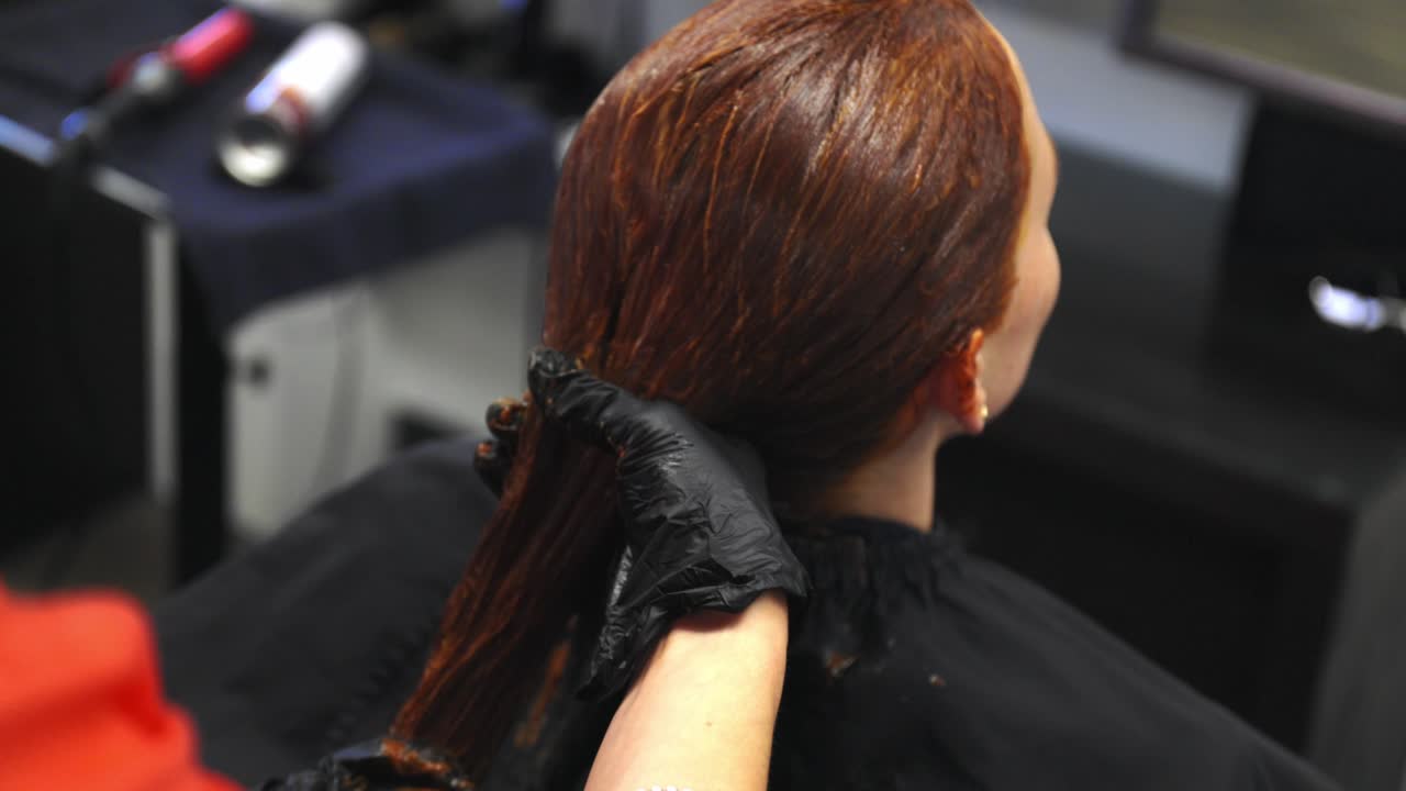 Parrucchiere/a con guanti medici neri che applica una tintura per capelli rossa a una donna caucasica seduta su una sedia da parrucchiere.
