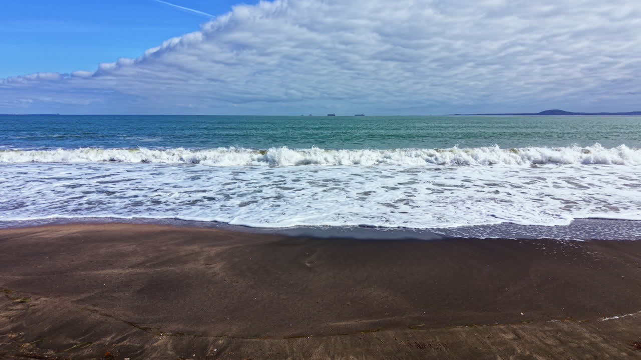 Vista delle onde dell'oceano che si infrangono sulla riva in una giornata di sole, con un ampio cielo pieno di nuvole su un orizzonte lontano.