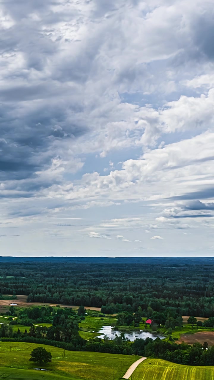 Aerial vertical timelapse of moving clouds above lone red roof house and private lake surrounded by dense forest, agricultural fields and meadows in green summer countryside