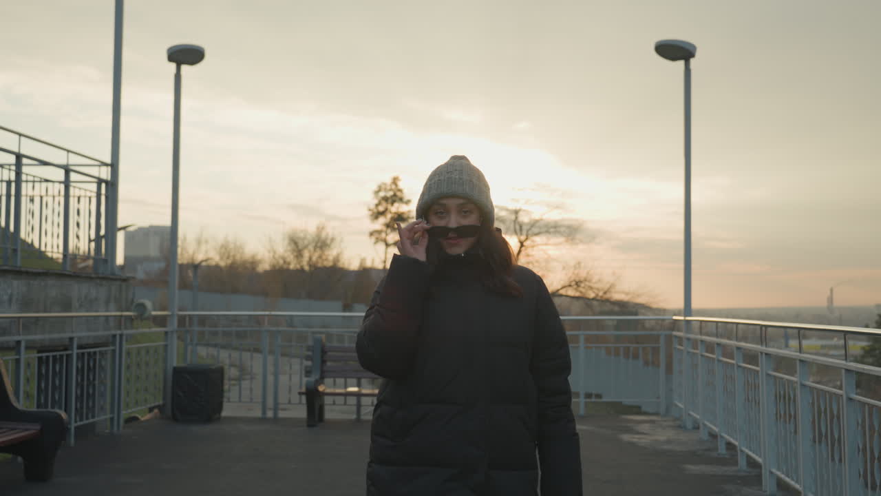 Solnedgang ved promenaden, kvinde går med solbriller, strikhue, vinterfrakke, bærer taske, varmt gyldent lys, rolig bysilhuet, blide skridt, kort smil mens hun taler i telefon, filmisk modlys, blødt.