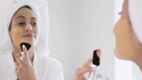 Happy-biracial-woman-applying-beauty-face-mask-looking-in-mirror-in-bathroom,-slow-motion