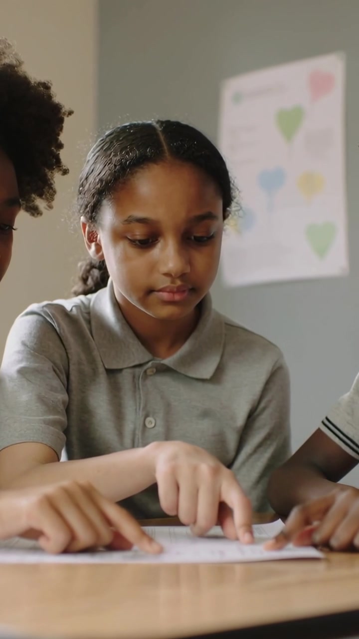 Vertical video: Leaning braided pupil in gray polo guiding index fingers tracing on paper at desk