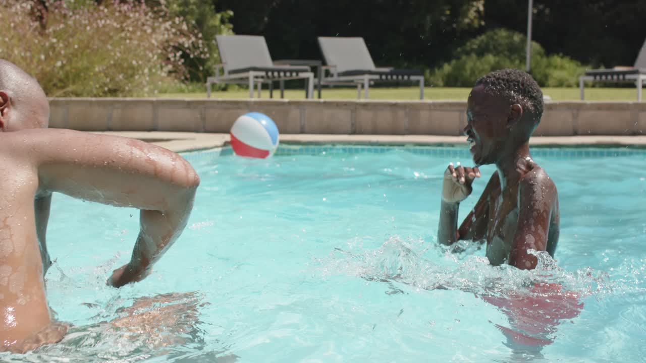 Homem afro-americano jogando uma bola de praia tricolor enquanto um adolescente espirra e brinca na piscina de casa.