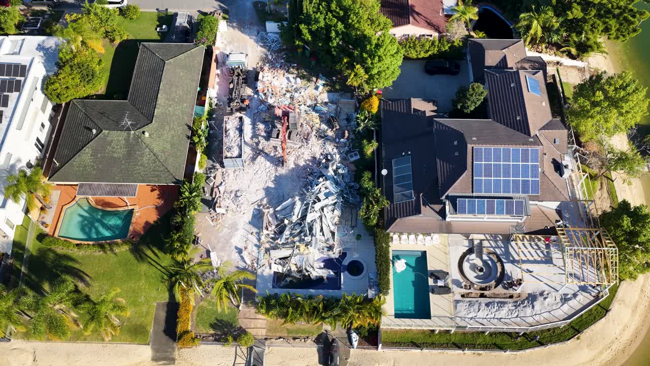 Drone footage captures a house being demolished by heavy machinery in a sunny suburban neighborhood, surrounded by modern homes with solar panels and swimming pools