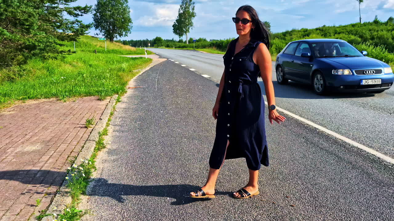 Donna in abito blu fa l'autostop a bordo strada mentre un'auto passa senza fermarsi.