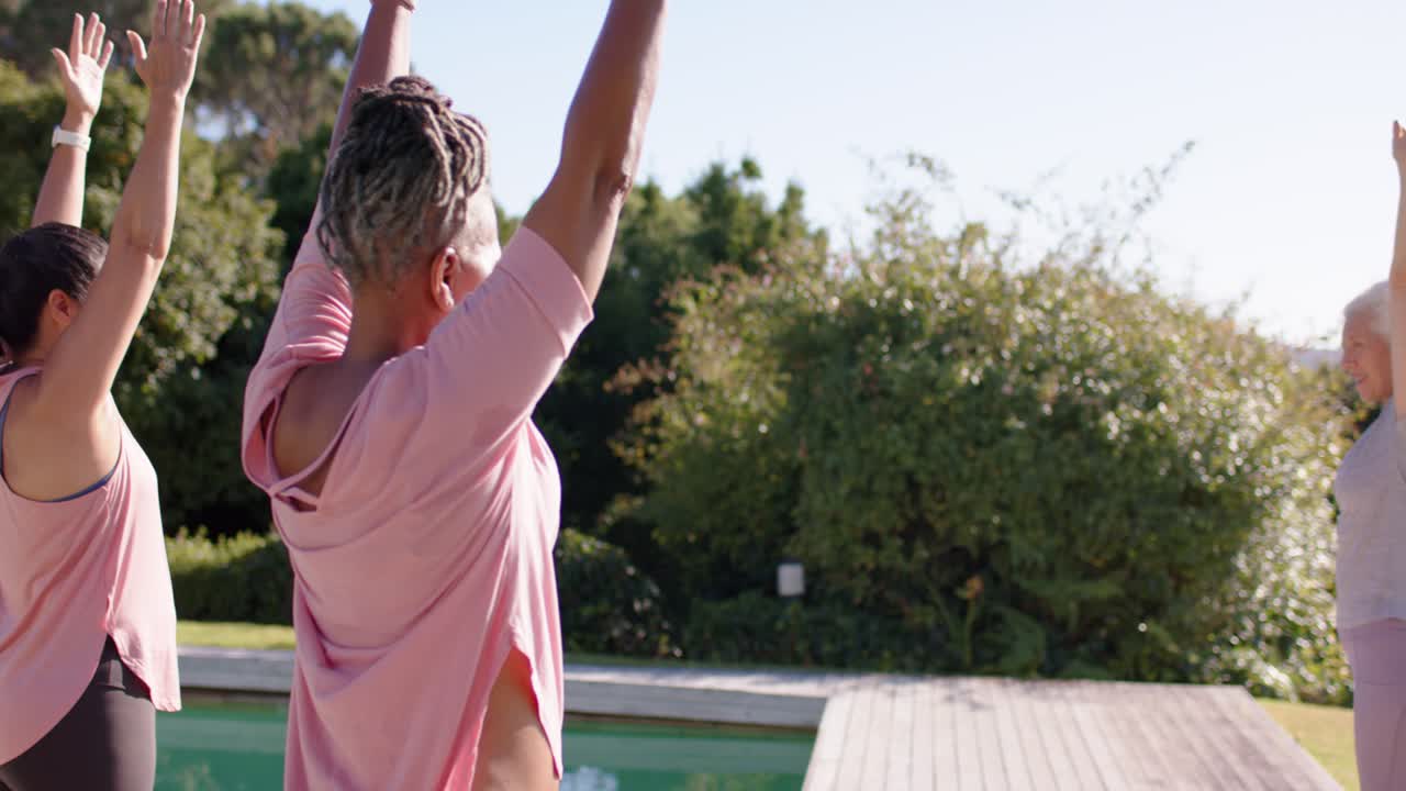 Diverse women mirroring senior leader raising arms, stretching overhead on deck with mat for warmup