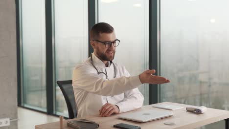 el médico con una bata de laboratorio sentado en un escritorio con una computadora portátil invita al paciente a sentarse frente a él