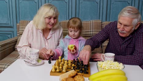 Mature grandmother grandfather with child girl grandchild playing chess game with on table in room